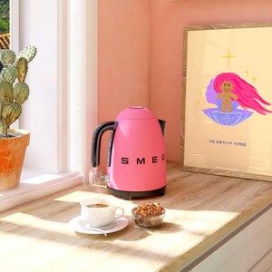 Op de afbeelding: Een roze elektrische waterkoker met een zwarte handgreep en tuit, het woord "SMEG" staat op de voorkant gedrukt, staat op een houten aanrechtblad naast een kopje koffie, een kom met ontbijtgranen en een ingelijst afdruk met een cartoonafbeelding van een rozeharige zeemeermin en de tekst "The Birth of Venus".