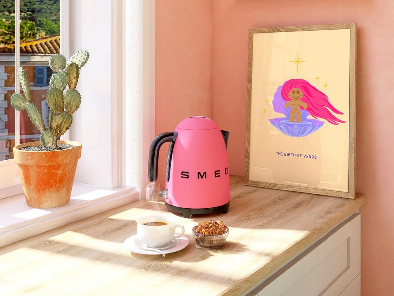 Op de afbeelding: Een roze elektrische waterkoker met een zwarte handgreep en tuit, het woord "SMEG" staat op de voorkant gedrukt, staat op een houten aanrechtblad naast een kopje koffie, een kom met ontbijtgranen en een ingelijst afdruk met een cartoonafbeelding van een rozeharige zeemeermin en de tekst "The Birth of Venus".
