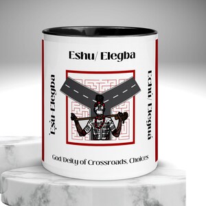 Puede incluir: Una taza de cerámica blanca con interior negro, con el texto "Eshu / Elegba" y "God Deity of Crossroads, Choices." El diseño incluye un gráfico de una figura sosteniendo un bastón en una encrucijada, con texto en rojo.