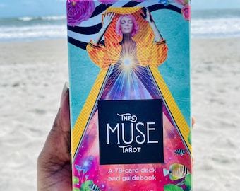 May include: A tarot card deck titled "The Muse Tarot" held in a hand. The box features a colorful illustration of a woman with pink hair, wearing an orange and yellow garment. The text "THE MUSE TAROT" is displayed on a black background, with the text "A 78-card deck and guidebook" below.