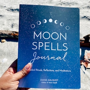May include: A journal with a dark blue cover featuring the words "MOON SPELLS Journal" in white script. The cover also displays a moon phase graphic and the author's name, Diane Ahlquist. The journal is held against a sandy background.
