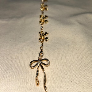 May include: A gold chain necklace with a bow charm and several smaller flower charms. The chain is delicate and the charms are intricate.