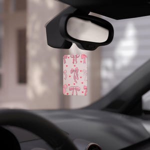 May include: Pink car air freshener with a pattern of pink bows and hearts on a white background.