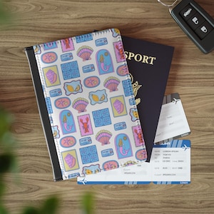 May include: A passport cover with a white background and a pattern of colorful sea creatures, shells, and mermaid illustrations. A dark blue passport and two boarding passes are visible on a wooden surface.