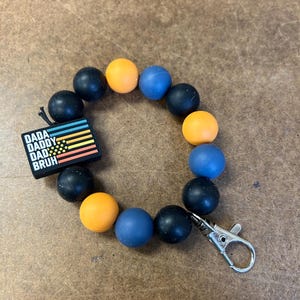 May include: A black, blue, and orange silicone beaded bracelet with a silver clasp. The bracelet has a charm with the text "DADA DADDY DAD BRUH" and a small flag design.