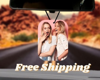May include: Two women are whispering to each other in a pink car air freshener. The women are smiling and looking at each other. The air freshener is hanging from the rearview mirror of a car.
