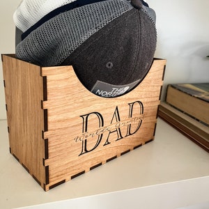 Könnte beinhalten: Hüte-Organizer aus Holz mit dem eingravierten Text "Dad World's Greatest" auf der Vorderseite. Der Organizer ist aus hellbraunem Holz gefertigt und hat einen dunkelbraunen Rand. Eine graue und weiße Baseballkappe liegt im Organizer.