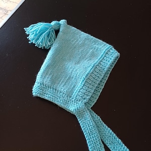 May include: A light blue knitted baby bonnet with a tassel and ties. The bonnet is made of soft, textured yarn. The ties can be tied under the chin. The bonnet is a light blue colour. The bonnet is a traditional style.