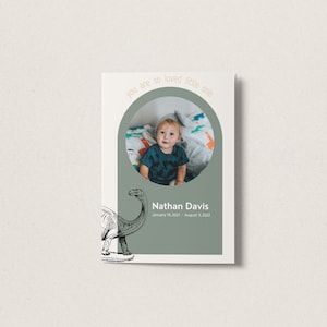 May include: A green and white card with a photo of a young boy in a circle. The text "You are so loved little one" is written in a peach color around the circle. Below the photo is the text "Nathan Davis" and "January 19, 2021 - August 1, 2022". A black and white illustration of a dinosaur is in the bottom left corner.