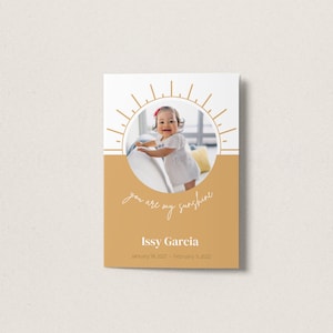 Peut inclure: Une carte blanche avec un motif de soleil doré et une photo d'une jeune fille. Le texte "you are my sunshine" est écrit en cursive sous la photo. La carte comprend également le nom "Issy Garcia" et les dates "January 18, 2021 - February 5, 2022".
