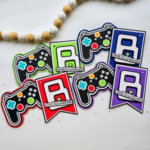 May include: A collection of gaming-themed party decorations featuring black video game controllers with colorful buttons and swallowtail banners in red, green, blue, and purple. Each banner displays a large white letter A and the text Level 10 Unlocked.