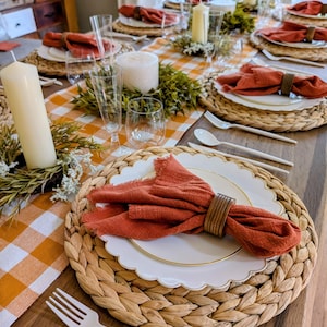 May include: A table setting featuring a woven round placemat, a white scalloped plate with a gold rim, and a rust-colored linen napkin secured with a wooden ring. The table includes a yellow and white checkered runner, white candles, and greenery.