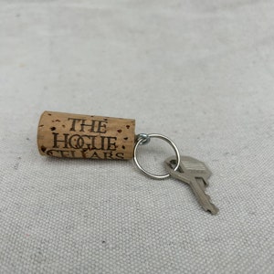 May include: A brown cork keychain with the text "THE HOGIE CELLARS" engraved on it. The keychain has a silver key ring with a silver key attached.
