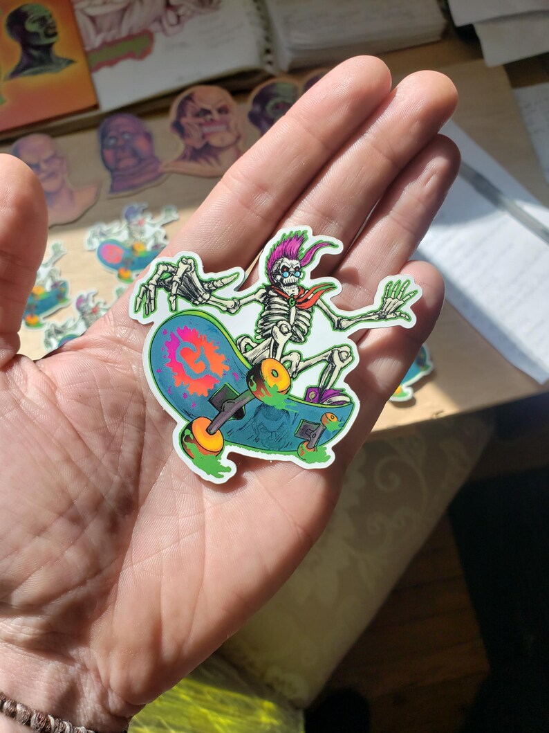 May include: A sticker of a skeleton skateboarding. The skeleton is wearing a pink and red bandana and has a green and blue skateboard with a green and yellow design.