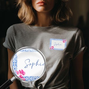 以下が含まれることがあります： 「Sophia」という名前が筆記体で書かれた長方形のデザインが特徴のグレーのTシャツ。 デザインは青と白のタイルのパターンとピンクの花柄のアクセントで縁取られています。 拡大鏡はデザインのクローズアップを示しています。