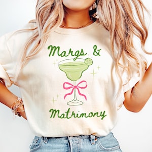 May include: Cream-colored t-shirt with the text "Margs & Matrimony" in green script. A green margarita glass with a lime wedge is centered above a pink bow. The shirt is a casual, fun design.