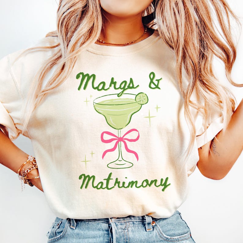 May include: Cream-colored t-shirt with the text "Margs & Matrimony" in green script. A green margarita glass with a lime wedge is centered above a pink bow. The shirt is a casual, fun design.