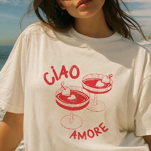 K&ouml;nnte beinhalten: Wei&szlig;es T-Shirt mit roter Grafik von zwei Cocktailgl&auml;sern mit herzf&ouml;rmigen Garnierungen. Der Text "CIAO AMORE" ist rot aufgedruckt. Das T-Shirt besteht aus einem weichen Material und ist f&uuml;r den Alltag geeignet.