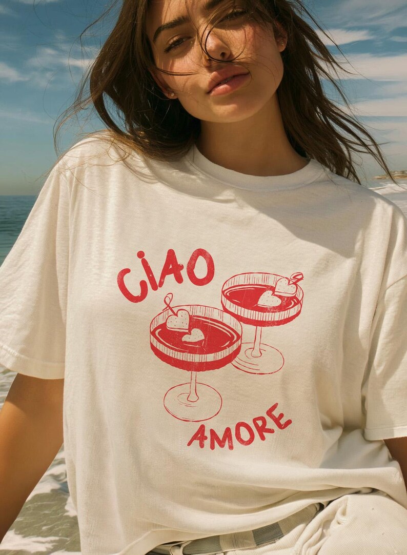 K&ouml;nnte beinhalten: Wei&szlig;es T-Shirt mit roter Grafik von zwei Cocktailgl&auml;sern mit herzf&ouml;rmigen Garnierungen. Der Text "CIAO AMORE" ist rot aufgedruckt. Das T-Shirt besteht aus einem weichen Material und ist f&uuml;r den Alltag geeignet.