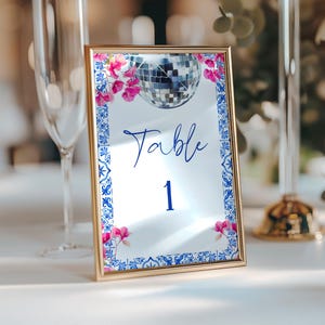 K&ouml;nnte beinhalten: Eine goldgerahmte Tischnummerkarte mit verspiegelter Oberfl&auml;che. Die Karte zeigt eine Discokugel-Grafik, rosa Blumenakzente und einen blau-wei&szlig;en Bord&uuml;renrand. Das Wort "Table" und die Zahl "1" sind in Blau geschrieben.