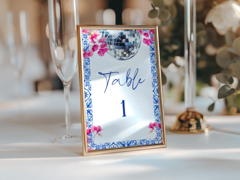K&ouml;nnte beinhalten: Eine goldgerahmte Tischnummerkarte mit verspiegelter Oberfl&auml;che. Die Karte zeigt eine Discokugel-Grafik, rosa Blumenakzente und einen blau-wei&szlig;en Bord&uuml;renrand. Das Wort "Table" und die Zahl "1" sind in Blau geschrieben.
