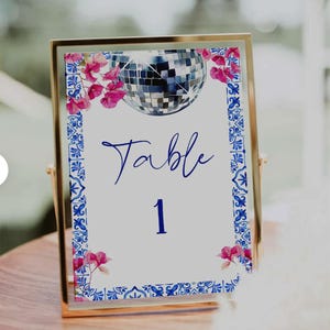 K&ouml;nnte beinhalten: Eine goldgerahmte Tischnummerkarte mit wei&szlig;em Hintergrund. Die Karte zeigt eine Discokugel-Illustration, rosa florale Akzente und blaue dekorative Bord&uuml;ren. Das Wort "Table" und die Zahl "1" sind in Blau geschrieben.