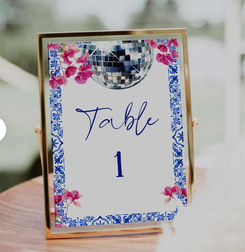 K&ouml;nnte beinhalten: Eine goldgerahmte Tischnummerkarte mit wei&szlig;em Hintergrund. Die Karte zeigt eine Discokugel-Illustration, rosa florale Akzente und blaue dekorative Bord&uuml;ren. Das Wort "Table" und die Zahl "1" sind in Blau geschrieben.