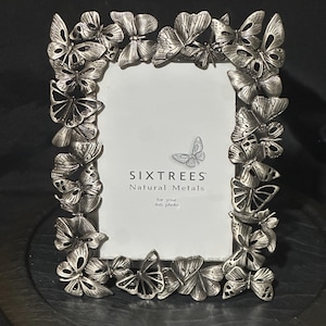 May include: A rectangular silver-toned metal picture frame decorated with numerous sculpted butterflies along the border. The center insert features the text SIXTREES Natural Metals and indicates it is for a 4x6 inch photo.