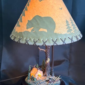 May include: A decorative lamp with a forest theme. The lampshade features a bear silhouette and trees. The base includes a miniature campsite with a tent and canoe. The lamp has a dark brown wooden base and a light-colored lampshade.