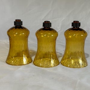 May include: Three amber-colored glass candle holders with a textured, dimpled surface. Each holder features a flared base, a curved body, and a small black plastic attachment at the top center, designed to hold a candle securely in place.