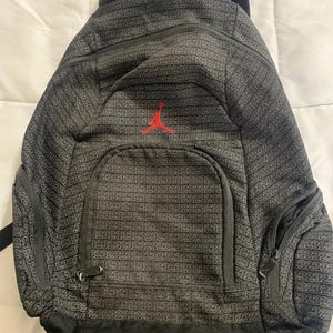 K&ouml;nnte beinhalten: Schwarzer Rucksack mit einem sich wiederholenden Muster der Zahl 23. Der Rucksack hat ein gro&szlig;es Hauptfach, eine kleinere Fronttasche und eine kleine Rei&szlig;verschlusstasche an der Seite. Die Vorderseite des Rucksacks zeigt ein rotes gesticktes Jumpman-Logo.