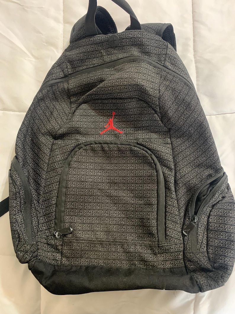 K&ouml;nnte beinhalten: Schwarzer Rucksack mit einem sich wiederholenden Muster der Zahl 23. Der Rucksack hat ein gro&szlig;es Hauptfach, eine kleinere Fronttasche und eine kleine Rei&szlig;verschlusstasche an der Seite. Die Vorderseite des Rucksacks zeigt ein rotes gesticktes Jumpman-Logo.