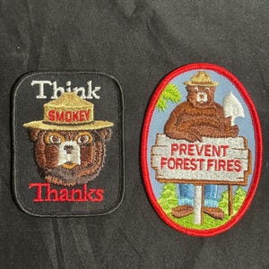 May include: Two embroidered patches featuring Smokey Bear. The left rectangular patch is black with the text Think and Thanks in white and red, showing a bear in a tan hat. The right oval patch has a red border, showing a bear holding a shovel.