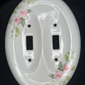 May include: An oval-shaped white ceramic double light switch wall plate featuring a floral pattern with pink flowers, green leaves, and small yellow buds. The plate has two rectangular cutouts for switches and four screw holes.