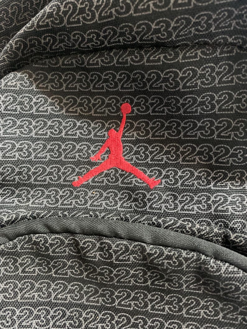 K&ouml;nnte beinhalten: Schwarzer Rucksack mit einem roten gestickten Basketballspieler-Logo und der Zahl 23, die sich in einem Muster auf dem Stoff wiederholt.