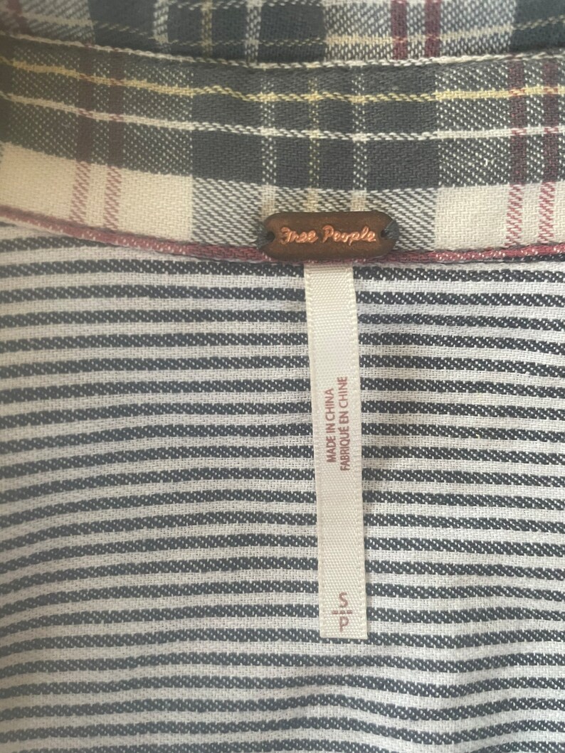 Pode incluir: Uma camisa listrada em preto e branco com uma etiqueta Free People. A etiqueta &eacute; marrom com texto branco e diz "Free People". A etiqueta tamb&eacute;m diz "Made in China" e "Fabriqu&eacute; en Chine".