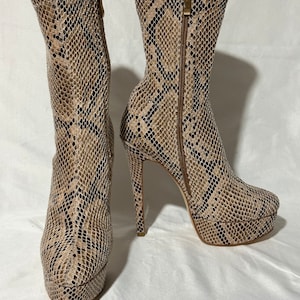 May include: A pair of snakeskin heeled ankle boots with a platform sole. The boots are a light brown color with a black snakeskin pattern.