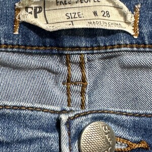 May include: A close-up of a blue denim jean with a Free People label that says "Size: W 28" and "Made in China". The jean has a silver button with the Free People logo.