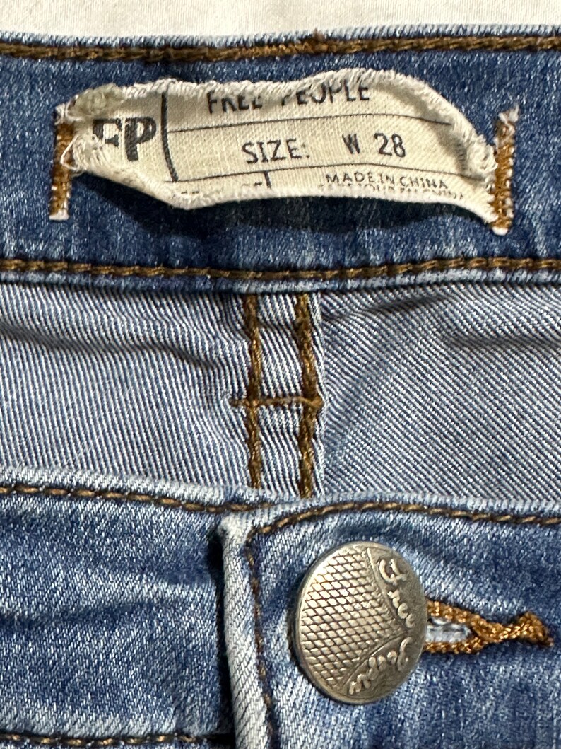 May include: A close-up of a blue denim jean with a Free People label that says "Size: W 28" and "Made in China". The jean has a silver button with the Free People logo.