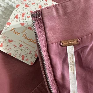 Op de afbeelding: Close-up van een stofroze rok met een zichtbare rits en een label van het merk "Free People". Het label heeft een bloemenpatroon en het logo van het merk. De rok heeft een gladde, leerachtige textuur.