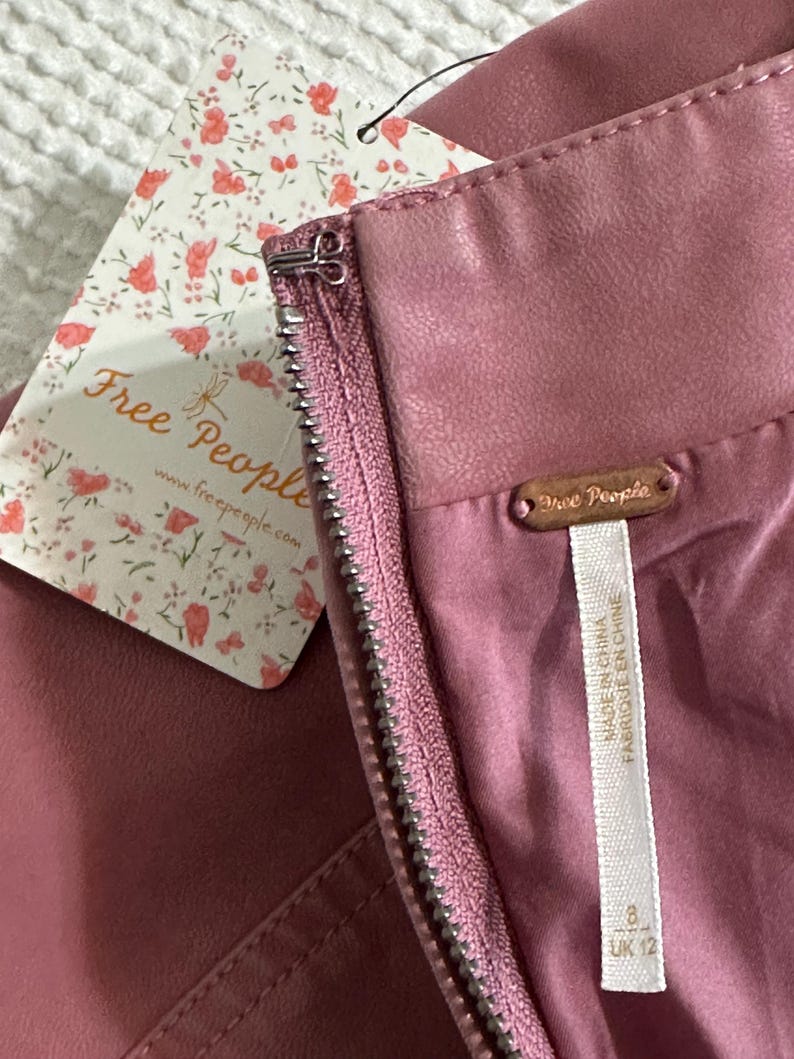 Op de afbeelding: Close-up van een stofroze rok met een zichtbare rits en een label van het merk "Free People". Het label heeft een bloemenpatroon en het logo van het merk. De rok heeft een gladde, leerachtige textuur.