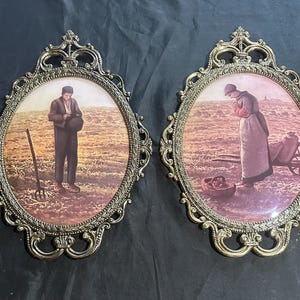 May include: Two oval wall plaques featuring ornate gold-toned metal frames with intricate scrollwork. Each plaque contains a print depicting a field scene: one shows a figure holding a hat in a field with a pitchfork, and the other shows a figure praying.