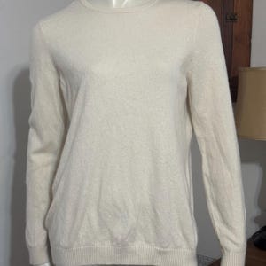 May include: A cream-colored, long-sleeved sweater with a crew neck. The sweater is displayed on a mannequin. The fabric appears soft and has a subtle texture. The cuffs and hem are ribbed.