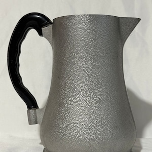 May include: A silver pitcher with a black plastic handle. The pitcher has a textured surface.