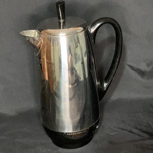 May include: A Farberware stainless steel coffee percolator with a black handle and base. The lid has a black knob. The percolator is reflective, showing the surrounding environment. The Farberware logo is visible on the base.