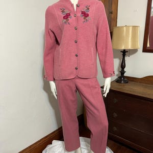 May include: A two-piece pink suit displayed on a mannequin. The jacket has a button-up front, two pockets, and floral embroidery. The matching pants are straight-legged. The suit is made of a textured fabric.
