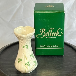 May include: A cream-colored Belleek Parian China vase with a textured surface and green shamrock motifs. The vase is accompanied by its original dark green rectangular box featuring the text Belleek Parian China Hand Crafted in Ireland.