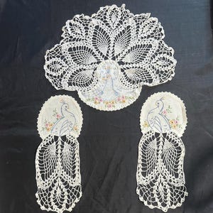 May include: A set of three off-white doilies featuring embroidered peacock motifs with blue bodies and colorful floral accents. The central doily has a large, fan-shaped crocheted tail, while the two smaller side doilies feature long, trailing crocheted tails.