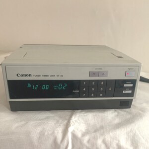 May include: A white Canon VT-20 tuner timer unit with a digital display showing 12:00 and 02. The unit has a black front panel with buttons for channel selection, charge, and timer.