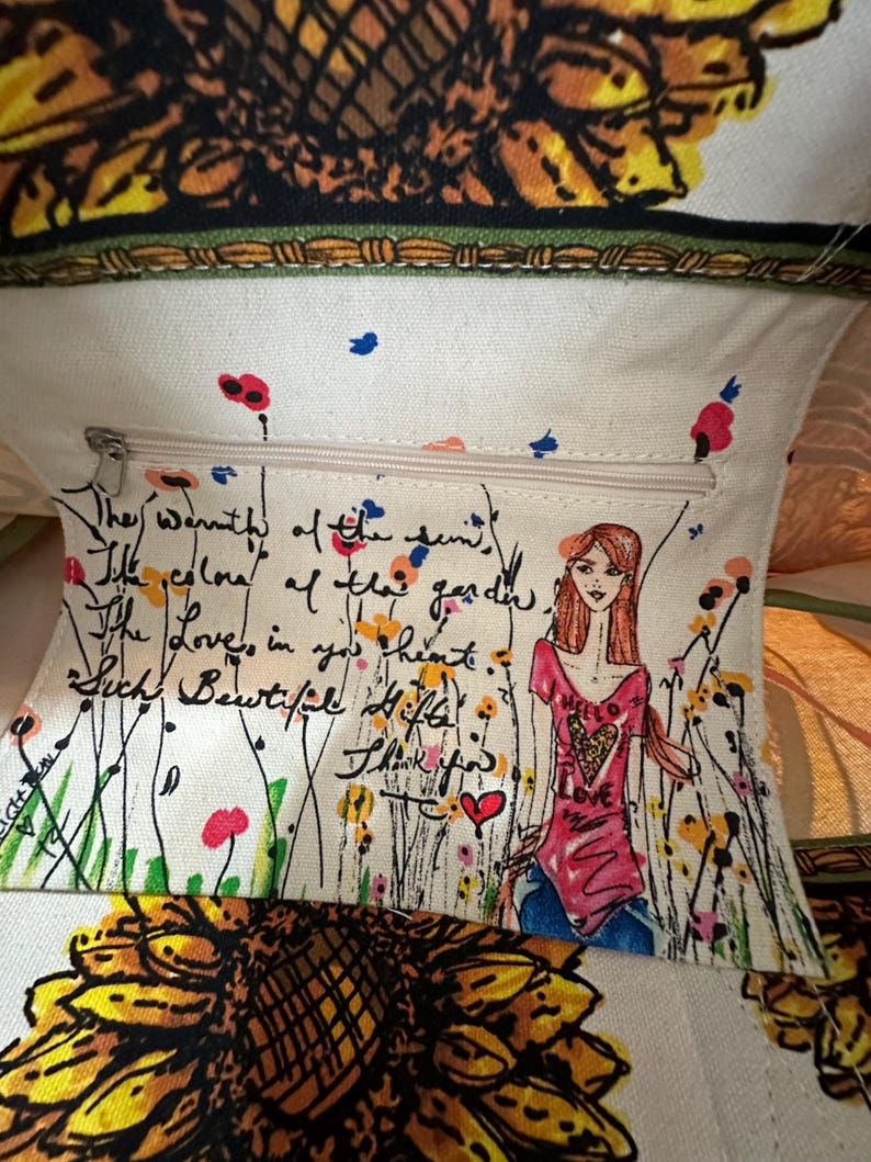 Peut inclure: Sac fourre-tout cr&egrave;me avec un motif de tournesol et un motif floral. L'int&eacute;rieur comporte une poche zipp&eacute;e avec un message manuscrit et un dessin d'une femme en chemise rose et jean bleu.